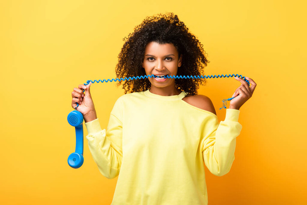curly african american woman biting cable of vintage telephone on yellow - Foto, afbeelding