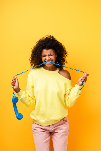 angry african american woman biting cable of vintage telephone on yellow - Photo, Image
