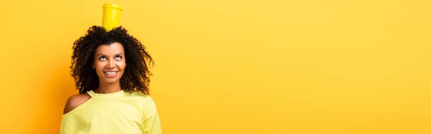 счастливая африканская американка с чашкой многоразового использования на голове на желтом, баннер - Фото, изображение