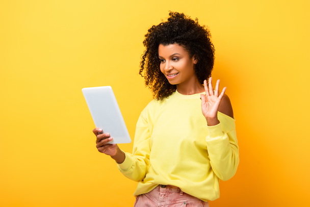 mujer afroamericana sosteniendo tableta digital y saludando de la mano mientras tiene video chat en amarillo - Foto, Imagen