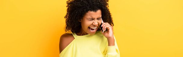 mujer afroamericana enojada sosteniendo teléfono inteligente y gritando en amarillo, pancarta - Foto, Imagen