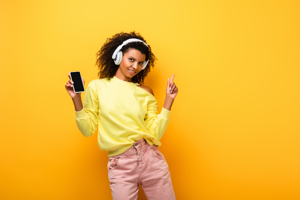 felice donna afroamericana in cuffia che tiene smartphone con schermo bianco mentre punta con il dito sul giallo - Foto, immagini