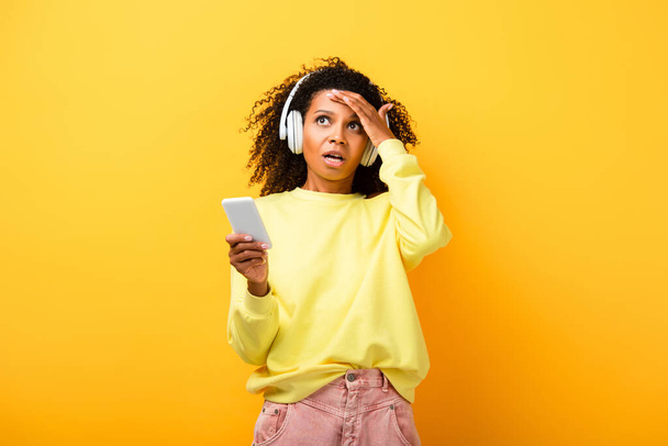 confused african american woman in headphones holding smartphone on yellow - Foto, immagini