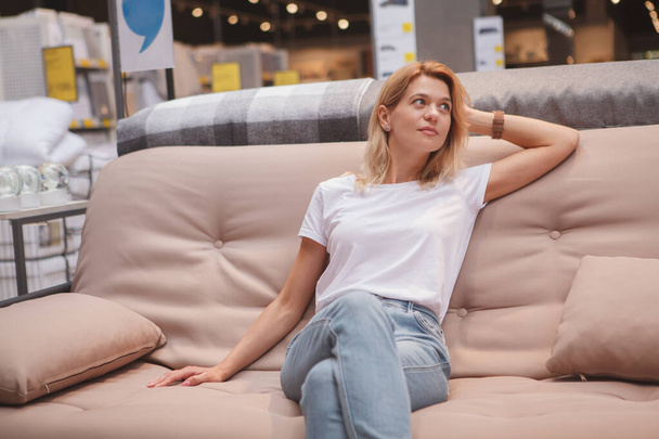 Encantadora mujer madura sentada en un sofá, mirando hacia otro lado, comprando muebles nuevos - Foto, Imagen