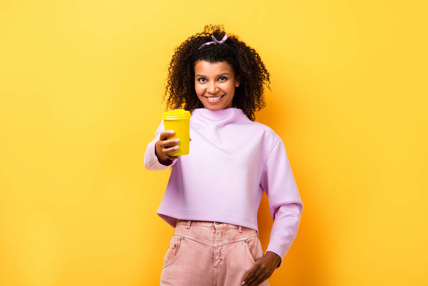 alegre mujer afroamericana sosteniendo la taza termo y de pie con la mano en el bolsillo en amarillo - Foto, Imagen