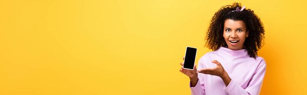 donna afroamericana eccitata che punta con mano lo smartphone con schermo bianco su giallo, banner - Foto, immagini