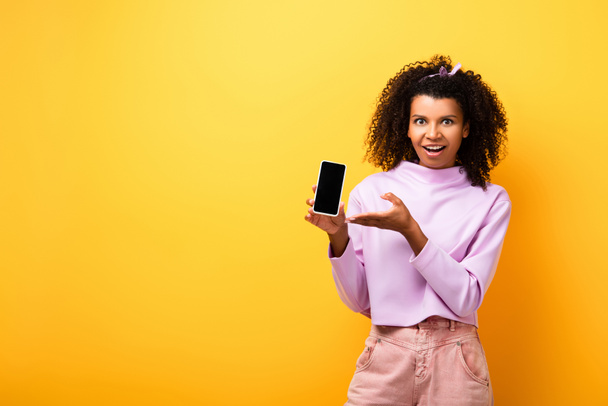 Heyecanlı Afrikalı Amerikalı kadın elinde akıllı telefonuyla sarı ekranda boş ekranı gösteriyor. - Fotoğraf, Görsel