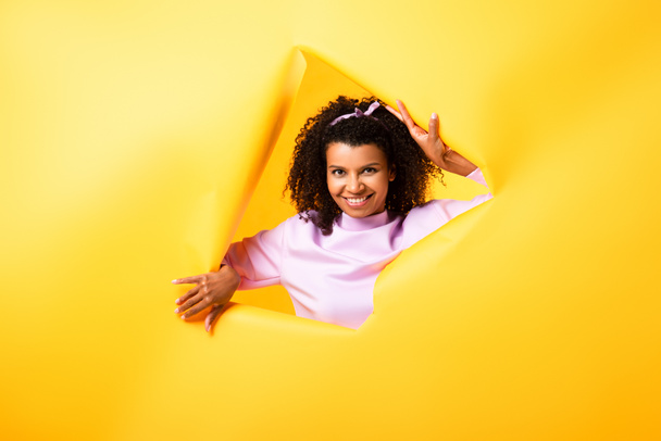happy african american woman looking at camera through hole in ripped paper on yellow background  - Foto, Bild