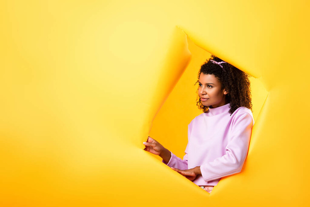Αφροαμερικανή γυναίκα κοιτάζει μακριά μέσα από την τρύπα σε σκισμένο χαρτί σε κίτρινο φόντο  - Φωτογραφία, εικόνα