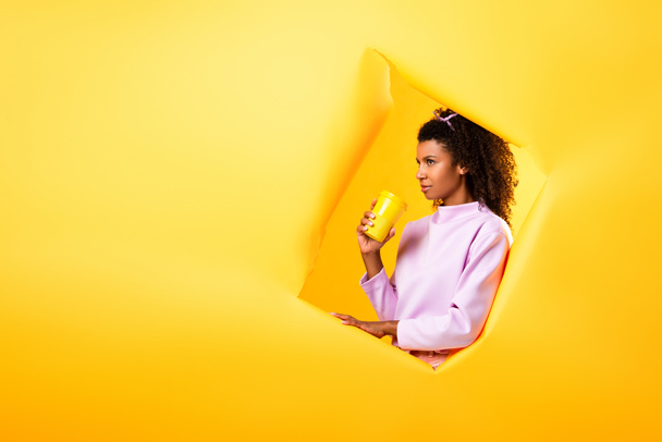 african american woman holding eco cup near hole in ripped paper on yellow background  - Fotoğraf, Görsel