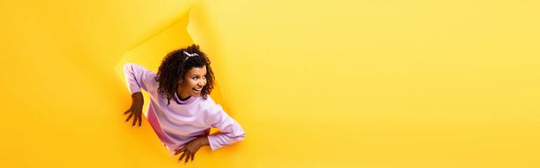 curious african american woman looking away through hole in ripped paper and smiling on yellow background, banner - Photo, Image