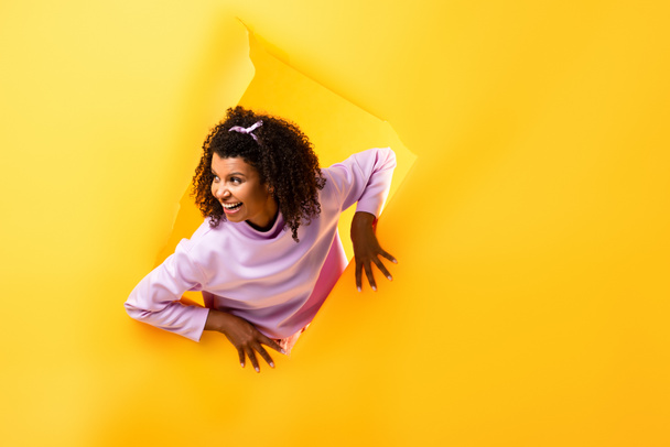 curiosa mulher afro-americana olhando para longe através do buraco em papel rasgado e sorrindo no fundo amarelo  - Foto, Imagem