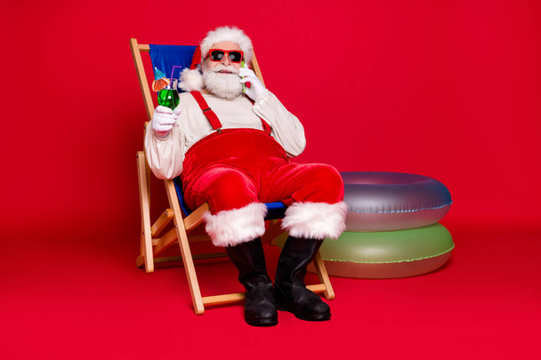 Porträt seiner schönen fröhlichen bärtigen dicken Weihnachtsmann Hipster sitzt im Stuhl trinkt Mojito Aufruf im Ausland Roaming Handy Ruhe entspannen isoliert hell lebendigen Glanz leuchtenden roten Hintergrund - Foto, Bild