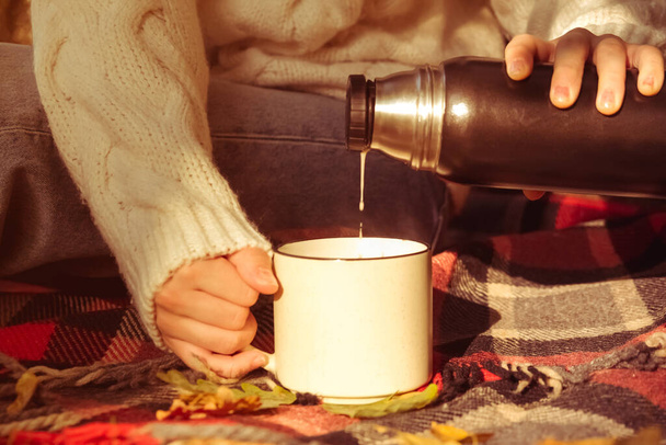 girl in a warm white sweater on a red plaid blanket pours coffee from a thermos. Autumn background, the concept of warmth and comfort - Фото, зображення