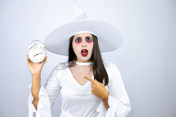 Woman wearing witch costume over isolated white background surprised, pointing and holding a clock - Photo, Image