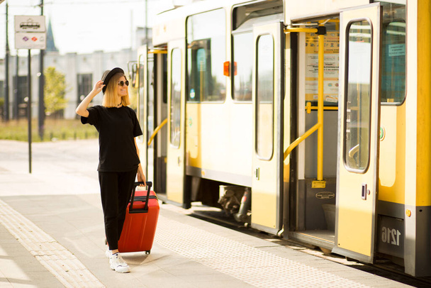 Стильна жінка в загальному чорному кольорі з червоною валізою на міській зупинці в очікуванні трамвая
 - Фото, зображення