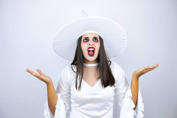 Donna che indossa costume da strega su sfondo bianco isolato espressione indiscreta e confusa con braccia e mani sollevate - Foto, immagini