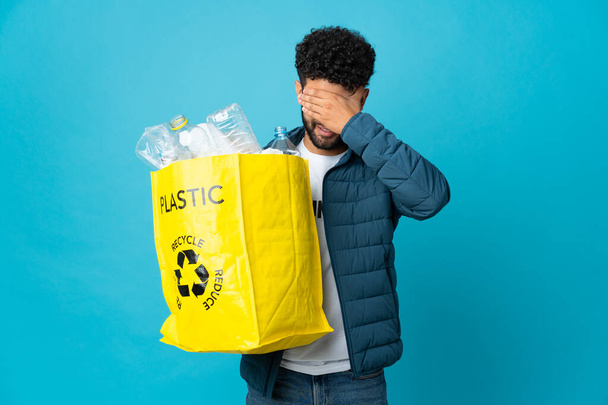 若いモロッコ人男性は、手で目を覆う孤立した背景にリサイクルするためにペットボトルでいっぱいの袋を持っています。何か見たくない - 写真・画像