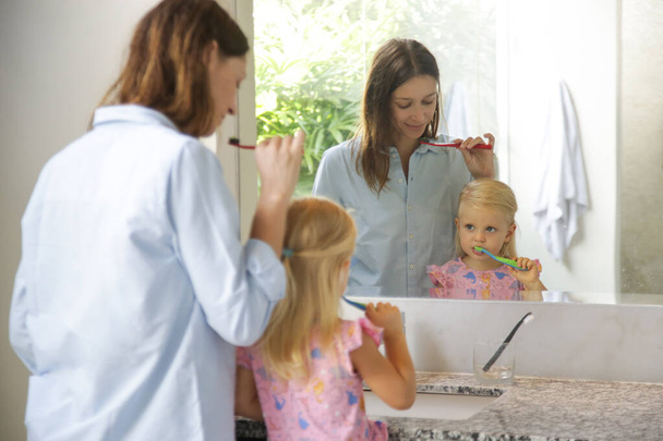 Η μητέρα και το τρίχρονο κορίτσι της βουρτσίζουν τα δόντια τους στο μπάνιο μπροστά στον καθρέφτη. - Φωτογραφία, εικόνα