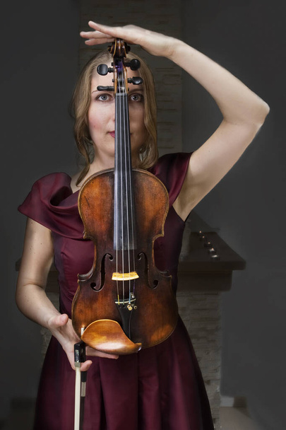 jonge violist in een fluwelen jurk houdt een meester viool in de buurt van de open haard, verticale indeling - Foto, afbeelding