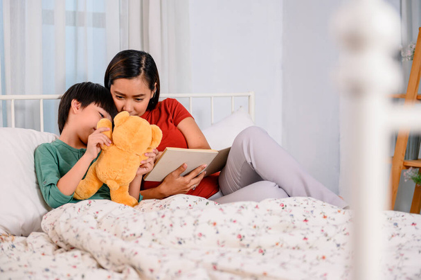Asian mother work home together with son. Mom and kid reading fairy tale before sleep on the bed. Woman lifestyle and family activity. - 写真・画像