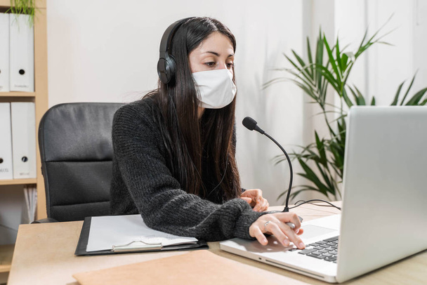 Telemarketer woman working on laptop and talking on a headset with face mask at office. Self employed and working from home because of coronavirus. Business worker answer customer calls.  - Photo, image