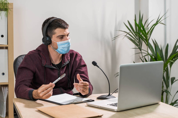 Telemarketer man working on laptop and talking on a headset with face mask at office. Self-isolated and working from home because of coronavirus. Business worker answer customer calls.  - Foto, afbeelding