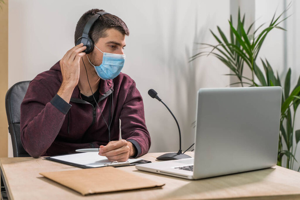 Telemarketer man working on laptop and talking on a headset with face mask at office. Self-isolated and working from home because of coronavirus. Business worker answer customer calls.  - Photo, image