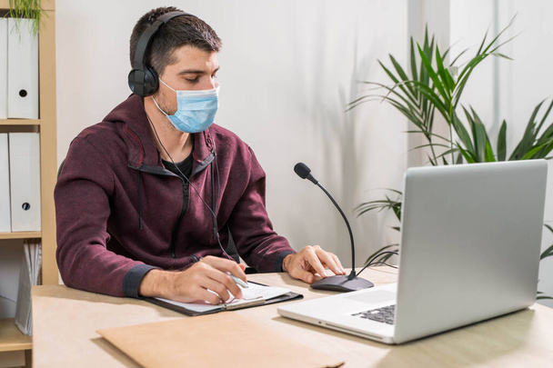 Telemarketer man working on laptop and talking on a headset with face mask at office. Self-isolated and working from home because of coronavirus. Business worker answer customer calls.  - Foto, afbeelding