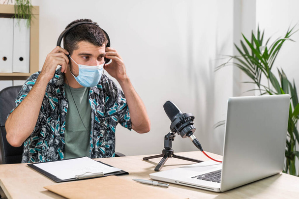 Man working as radio host at radio station sitting in front of microphone. Young radio host moderating a live show for radio with face mask. Working at home. Coronavirus. - Фото, зображення