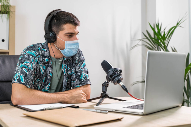 Man working as radio host at radio station sitting in front of microphone. Young radio host moderating a live show for radio with face mask. Working at home. Coronavirus. - Foto, afbeelding