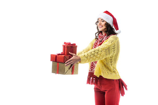 joyful young woman in santa hat, scarf and knitted sweater holding gifts isolated on white - Foto, Imagem