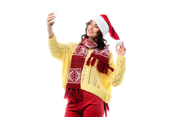 joyful woman in santa hat and scarf taking selfie on smartphone isolated on white  - Photo, Image