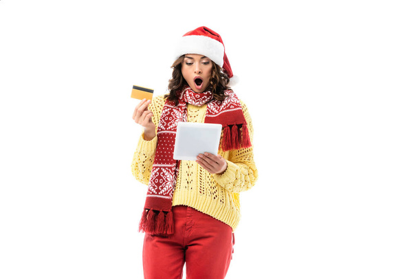 Şok olmuş genç bir kadın Noel Baba şapkası ve eşarbıyla dijital tablete bakarken elinde beyaz bir kart tutuyor.  - Fotoğraf, Görsel