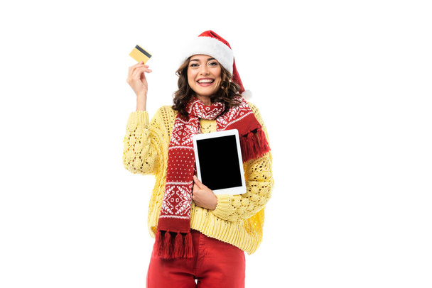 freudige Frau mit Weihnachtsmütze und Schal, die ein digitales Tablet mit leerem Bildschirm und Kreditkarte auf weißem Hintergrund hält  - Foto, Bild