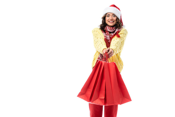 fröhliche junge Frau mit Weihnachtsmütze und Schal mit Ornament, die rote Einkaufstüten auf weißem Grund hält  - Foto, Bild