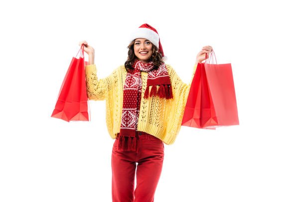pleased woman in santa hat and scarf with ornament holding red shopping bags isolated on white  - 写真・画像