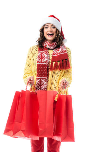 izgatott fiatal nő télapó kalap és sál dísz gazdaság piros bevásárló táskák elszigetelt fehér  - Fotó, kép