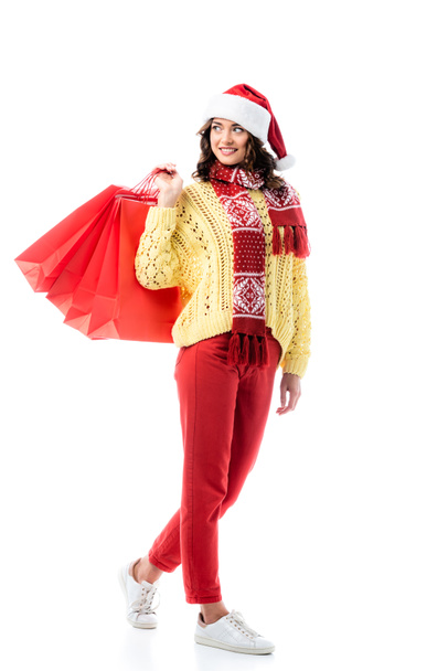 donna soddisfatta in cappello di Babbo Natale e sciarpa con ornamento tenendo borse della spesa isolate su bianco  - Foto, immagini
