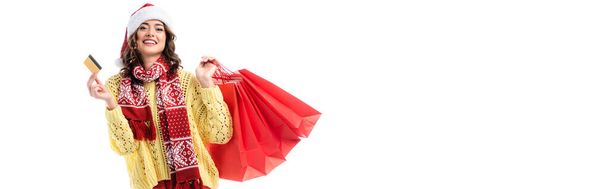 concept panoramique de femme heureuse en santa chapeau et écharpe avec ornement tenant sacs à provisions et carte de crédit isolé sur blanc  - Photo, image