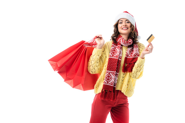 freudige Frau in Weihnachtsmütze und Schal mit Ornament, die Einkaufstaschen und Kreditkarte isoliert auf weiß hält  - Foto, Bild