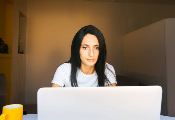 close-up portret van jonge blanke vrouw op zoek recht vooruit naar laptop in gezellige woonkamer - Foto, afbeelding