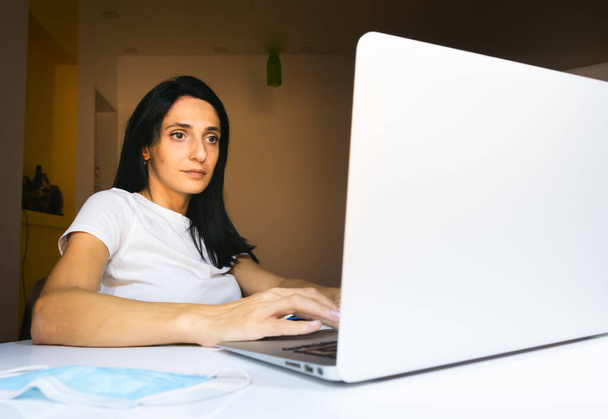Junge kaukasische Frau sitzt in Wohnung hinter weißem Schreibtisch und blickt geradeaus. Arbeitskonzept von zu Hause aus. - Foto, Bild