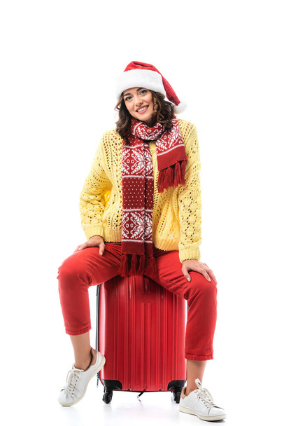 vreugdevolle jonge vrouw in santa hoed en sjaal met ornament zitten op reistas geïsoleerd op wit - Foto, afbeelding