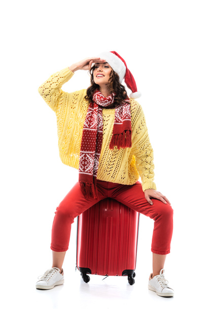 freudige junge Frau in Weihnachtsmütze und Schal mit Ornament sitzt auf Reisetasche und schaut isoliert auf weißem Grund weg - Foto, Bild