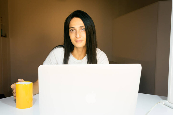 Portatif bilgisayarın arkasında genç beyaz bir kadın kameraya bakıyor ve elinde kocaman sarı bir kupa var. - Fotoğraf, Görsel