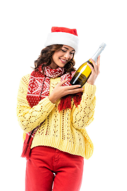 freudige junge Frau mit Weihnachtsmütze und Schal mit Ornament, die auf eine Champagnerflasche auf weißem Grund blickt  - Foto, Bild