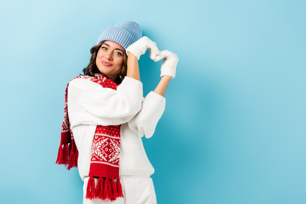 pleased young woman in winter outfit, warm scarf, gloves and hat showing heart sign on blue - Фото, зображення