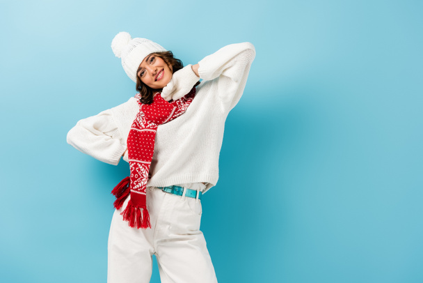 pleased young woman in white winter outfit, warm scarf, gloves and hat on blue  - Foto, immagini