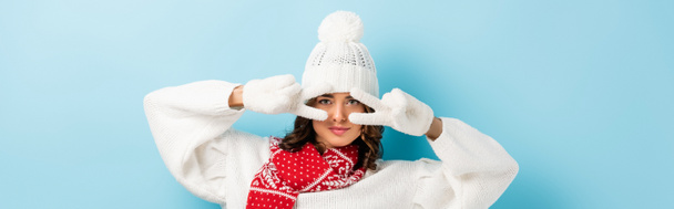 panoramic crop of young woman in white winter outfit showing peace sign on blue  - Photo, Image